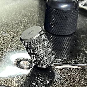 Puede incluir: Primer plano de dos perillas de guitarra negras con superficies texturizadas. Una perilla está enfocada, mostrando crestas detalladas y una parte superior plateada. La otra perilla está ligeramente desenfocada, con un diseño texturizado similar. Las perillas están sobre una superficie oscura y reflectante.