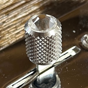 Puede incluir: Un pomo plateado con una parte superior en forma de diamante. El pomo tiene un patrón texturizado en forma de diamante.