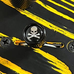 May include: Close-up of a black guitar with yellow stripes and a skull and crossbones knob. The knob is centered, with two screws visible on either side. The design has a grunge aesthetic.