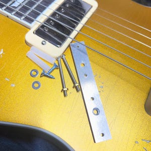 May include: Close-up of a gold electric guitar with a cream-colored pickup and silver strings. A silver metal plate, screws, washers, and plastic anchors are next to the guitar, suggesting repair or modification.