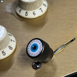 Puede incluir: Una perilla negra con un ojo blanco y venas rojas. La perilla está unida a un eje plateado.