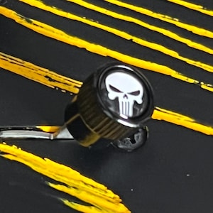 May include: A black gear shift knob featuring a white skull emblem. The knob is set against a black background with yellow diagonal stripes, creating a striking contrast. The skull design is a prominent detail.