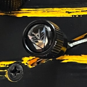 May include: Close-up of a black knob with a silver and black emblem. The emblem is a stylized face. The knob is set against a black background with yellow and orange paint strokes and a silver screw.