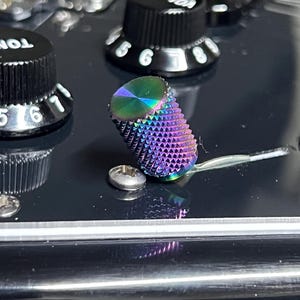 Może przedstawiać: Zbliżenie pokręteł gitary. Ostrość skupia się na tęczowym pokrętle głośności o teksturowanej, żłobionej powierzchni. Widoczne są również inne czarne pokrętła z białymi cyframi oraz srebrna śruba.