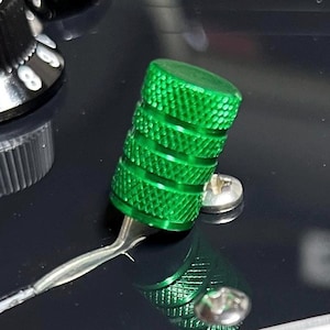Puede incluir: Un botón de control cilíndrico verde brillante con una superficie texturizada, probablemente para una guitarra u otro dispositivo electrónico. El botón está conectado a un cable plateado y se encuentra sobre una superficie negra, con un tornillo plateado visible. El diseño sugiere instrumentos musicales.