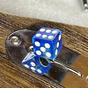 以下が含まれることがあります： 各面に白い点がついた青い透明なサイコロ。サイコロは銀色の金属製の表面の上に置かれています。