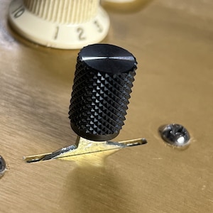 Puede incluir: Una perilla negra con un patrón de diamante en una superficie de metal dorado. La perilla está unida a una palanca de metal.