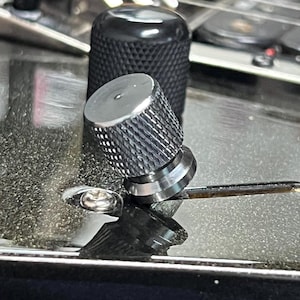 Puede incluir: Primer plano de las clavijas de afinación de una guitarra. Las clavijas son plateadas y negras, con un agarre texturizado para facilitar el giro. El cuerpo de la guitarra es visible en el fondo, reflejando la luz.