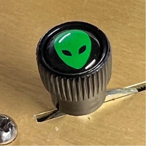 May include: A black knob with a green alien face graphic. The knob has a ridged edge.