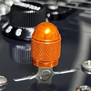 Puede incluir: Primer plano de un botón de guitarra de metal naranja con un agarre texturizado y una parte superior redondeada. Un botón de volumen negro está en el fondo, con los números 5 y 6 visibles. La superficie de la guitarra es oscura.