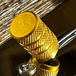 Op de afbeelding: Close-up van een goudkleurige, getextureerde knop met een ruitpatroon. De knop heeft een cilindrische vorm met een platte bovenkant en is geplaatst tegen een donker, reflecterend oppervlak. De knop is waarschijnlijk een decoratief of functioneel hardware-element.