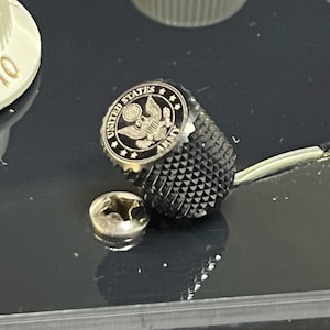 May include: A black metal guitar control knob with a knurled texture and a circular top insert featuring the United States Army emblem, including an eagle and the text UNITED STATES ARMY, positioned next to a silver Phillips head mounting screw.