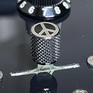 Puede incluir: Primer plano de un botón de guitarra negro con un símbolo de la paz. El botón tiene una superficie texturizada y está sobre un fondo negro. Los números 5, 6, 7, 8 y 9 son visibles en un botón cercano.