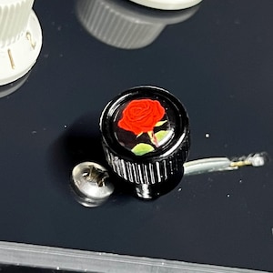 May include: A black cylindrical guitar control knob featuring a glossy red rose graphic on its top surface. The knob has a ribbed side texture for grip and is positioned next to a small silver metal screw on a smooth, dark, reflective surface.