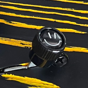 May include: A black knob with a smiley face design. The knob is set against a black background with yellow horizontal stripes. The knob has a screw on the side.