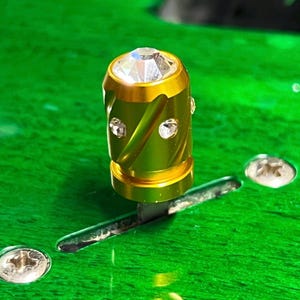 Puede incluir: Un selector de pastillas cilíndrico de metal dorado para guitarra, con un cristal transparente facetado en la parte superior y cristales más pequeños incrustados en los laterales, montado sobre un cuerpo de guitarra verde brillante con tornillos plateados.