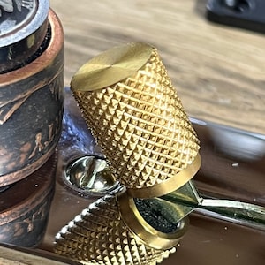 May include: A gold-coloured metal knob with a diamond-shaped pattern. The knob is attached to a metal surface.