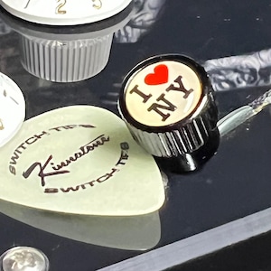 Peut inclure: Un bouton de contrôle de guitare noir avec une incrustation circulaire blanche ornée d'une icône de cœur rouge et du texte noir I NY, placé à côté d'un médiator vert pâle portant l'inscription noire Kinnatone Switch Tips sur sa surface.