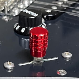 Puede incluir: Primer plano del panel de control de una guitarra con un botón rojo y un botón negro con la palabra "VOLUME" y números. El panel es negro, con tornillos plateados y una placa metálica.