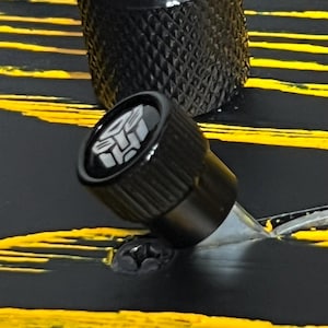 May include: Close-up of a black knob with a silver stem and a logo on the top. The knob is set against a black and yellow striped background. The top knob is also black and textured.