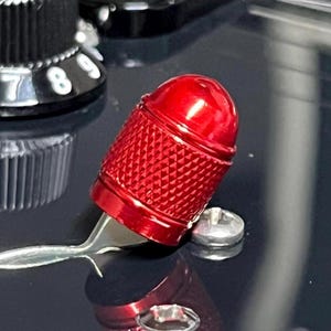 Puede incluir: Primer plano de un botón rojo texturizado con base plateada. El botón es cilíndrico con la parte superior redondeada y un patrón de diamantes. El fondo es una superficie oscura y reflectante con partes de otros objetos visibles.