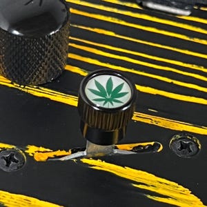 May include: Close-up of a black guitar knob featuring a green cannabis leaf design. The knob is set against a black surface with yellow stripes. Another black knob and screws are also visible.