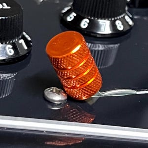 Könnte beinhalten: Nahaufnahme eines leuchtend orangefarbenen Gitarrenknopfs mit einem strukturierten, geriffelten Design. Der Knopf ist zylindrisch und sitzt auf einer schwarzen Oberfläche, in der Nähe anderer schwarzer Knöpfe mit weißen Zahlenmarkierungen. Eine kleine silberne Schraube ist ebenfalls sichtbar.