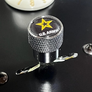 Puede incluir: Una perilla plateada con fondo negro y una estrella amarilla con el texto "U.S. ARMY" en ella.