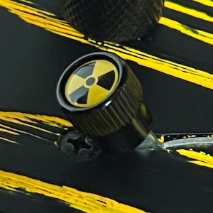 以下が含まれることがあります： 黄色と黒の放射線マークが付いた黒いノブのクローズアップ。ノブは、黄色の斜めストライプが付いた黒い表面に配置されています。デザインは危険または警告のテーマを示唆しています。