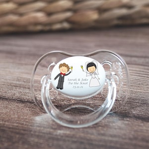 Pode incluir: Uma chupeta branca com um adesivo redondo branco que mostra um desenho animado de uma noiva e um noivo segurando taças de champanhe. O adesivo diz "Sarah & Jake Tie the Knot 23.11.21".