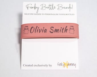 Könnte beinhalten: Ein rosa Silikon-Flaschenband mit dem Text "Olivia Smith" in schwarzer Schrift. Das Band zeigt ein kleines Cartoon-Bild einer Person mit einem Schnuller. Die Texte "Funky Bottle Bands!" und "Exklusiv kreiert von funky dummy" sind auf dem weißen Hintergrund gedruckt.