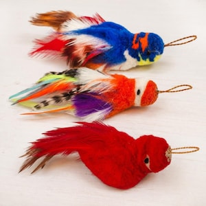 May include: Three decorative bird ornaments crafted from felt and feathers. The top bird features blue, white, and red plumage. The middle bird displays orange, white, purple, and striped feathers. The bottom bird is solid red with brown tail feathers.