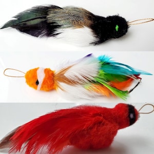 May include: Three colorful cat toys with feathers. The first toy is black and white with green eyes. The second toy is orange and white with a fish design. The third toy is red with black eyes.