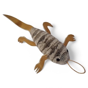 Peut inclure: Un jouet en peluche en forme de lézard avec un corps rayé marron et gris, des pattes et une queue en simili cuir marron et de petits yeux noirs. Une boucle de ficelle s'étend de la bouche du lézard. Le jouet est conçu pour les animaux de compagnie.
