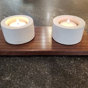 May include: Two white concrete candle holders with lit tea lights, sitting on a dark brown wooden tray.