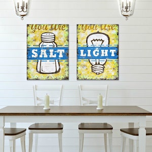 May include: Two framed prints with a yellow background and blue accents. The left print features a salt shaker with the text "You are Salt". The right print features a light bulb with the text "You are Light".