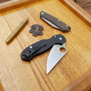 Könnte beinhalten: Ein Holztablett mit einem goldenen Stift, einer kleinen Kupfermünze mit einem Design, einem schwarzen und grünen Taschenmesser und einem schwarzen Taschenmesser mit einer silbernen Klinge.