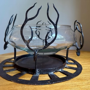 May include: A clear glass bowl sits in a decorative wrought iron stand. The stand has a circular base with cut-out designs and curved, antler-like supports that cradle the bowl. The metal has a dark, aged finish.