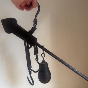 May include: Antique black metal balance scale with a hook, a weight, and a long arm. The scale is held by a hand. The scale is likely used for measuring weight. The scale is made of iron and has a rustic appearance.