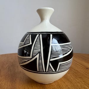 May include: A ceramic vase with a cream-colored top and a rounded black base. The black base features a geometric design of white lines and shapes. The vase has a narrow neck and a bulbous body, suitable for home decor.