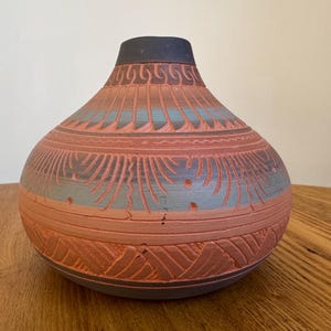 May include: A rounded terracotta vase featuring a dark charcoal neck and base. The surface is decorated with intricate, carved geometric patterns and horizontal bands in alternating shades of muted blue-gray and natural reddish-brown clay.