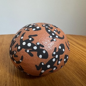 May include: A round, terracotta-colored decorative stone featuring a pattern of black lizards with white polka dots. The lizards are stylized and evenly spaced across the surface of the stone.