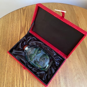 May include: A small, clear glass bottle with a red stopper and a painted scene inside. The bottle is in a red velvet box with a black satin lining.
