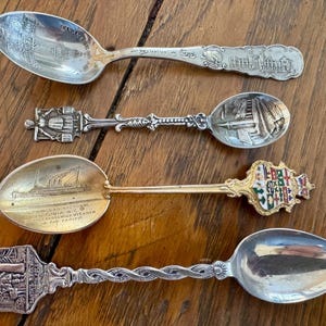 May include: Four antique souvenir spoons with detailed designs. The spoons are silver-toned, with intricate patterns on the handles and bowls. One spoon features a building, another a ship, and a third has a colorful crest. The spoons are displayed on a wooden surface.