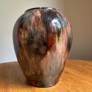 May include: A bulbous ceramic vase with a glossy finish featuring a marbled glaze pattern in shades of deep brown, black, cream, and rust orange. The vessel has a wide, rounded body that tapers slightly toward a circular opening at the top.