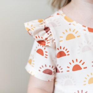 May include: A white short-sleeved shirt with a ruffled sleeve and a pattern of orange, yellow, and red suns.