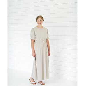 May include: A floor-length beige dress with short puff sleeves, a round neckline, and a cinched waistline. The dress features a simple, solid-colored fabric design that extends to the ankles, paired with white open-toe sandals with thin straps.