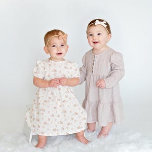 May include: Two babies wearing dresses and headbands. One baby is wearing a white dress with a floral pattern and a brown headband. The other baby is wearing a light pink dress and a white headband.