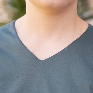 May include: Close-up of a teal-colored V-neck top. The fabric appears to be a soft, matte material. The neckline is a sharp V-shape, and the overall style is simple and elegant.