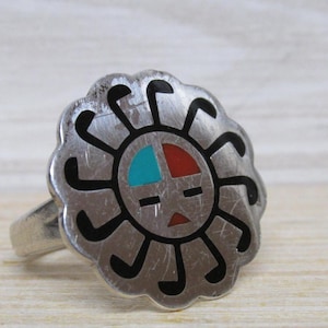 May include: A silver-toned ring with a sunburst design. The ring features a central sun face with turquoise and red accents, surrounded by black curved lines. The ring's band is a simple, polished silver.